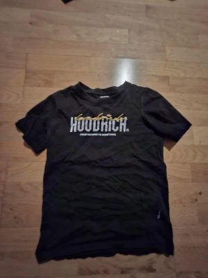 Svart Hoodrich t-shirt med tryck - Streetwear t-shirt från Hoodrich i svart med vit och gul logotyp på bröstet och coolt tryck på ryggen. Klassisk rund hals och korta ärmar. Materialet är mjuk bomull som känns skön mot huden. Perfekt för dig som gillar urban stil och snygga detaljer.