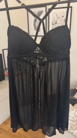 Svart spetsklänning Marilyn Monroe S - Svart kort klänning från Marilyn Monroe med vadderad byst, spetsdetaljer och tunna axelband som korsas i ryggen. Kjoldelen är transparent och luftig, vilket ger en riktigt snygg och feminin look. Perfekt för dig som gillar en edgy och chic stil.