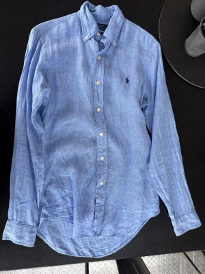 Blå linneskjorta från Polo Ralph Lauren - Använd ett par gånger. Snygg blå skjorta i linne från Polo Ralph Lauren med klassisk button down-krage och broderad logga på bröstet. Skjortan har långa ärmar, vita knappar och en avslappnad passform. Perfekt för en stilren och chill look.