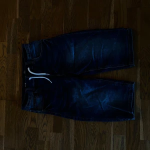 Blå denimshorts från LAB Industries - Säljer ett par blå denimshorts från LAB Industries med vit snörning i midjan och klassiska fem fickor. Shortsen har en loose passform och är tillverkade i bomull. Perfekta för dig som gillar streetwear och vill ha en chill look.