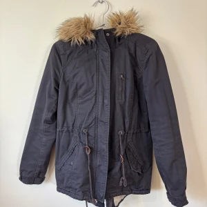  Parkasjacka - 36 Storlek - Svart parkasjacka med mysig beige fuskpäls i huvan och på insidan. Jackan har dragsko i midjan, flera fickor och stängs med dragkedja och knappar. 