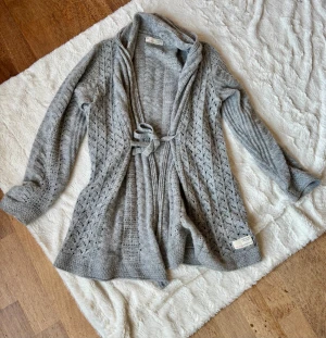 Grå stickad kofta Odd Molly med knyt - Superfin grå stickad kofta från Odd Molly med snyggt hålmönster och knytband i midjan. Långärmad och med mjuk, mysig känsla. Perfekt att slänga på sig när det är lite kyligt ute. Märkeslapp nertill och vid kragen.