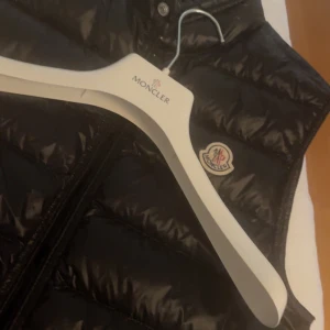 Svart dunväst Moncler Gui Gilet - Svart Moncler Gui Gilet dunväst med glansig finish och klassisk Moncler-logga på bröstet. Västen har en liten defekt (bild 4) som knappt syns annars är den i bra skick och jag säljer den för den är för liten. Kom dm för fler bilder och prisförslag!