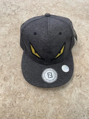 Grå keps från Fendi med gula ögon - Säljer en grå keps från Fendi med broderade gula ögon framtill och Fendi-logga baktill. Kepsen har klassisk snapback-modell och är tillverkad i ett mjukt bomullstyg. Perfekt för dig som vill sticka ut med en cool detalj.