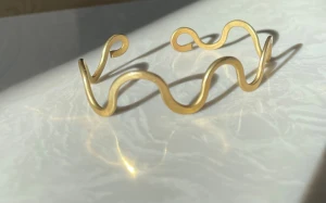 Vågigt guldarmband i aluminium  - Unikt armband i guldtonad metall med vågig, öppen design. Armbandet har en cool och modern look med mjuka, böljande former som ger en lekfull känsla. Perfekt för dig som vill sticka ut med ett stilrent men ändå annorlunda smycke.