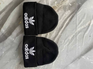 Svart Adidas mössa med broderad logga - Två svarta mössor från Adidas med klassisk vit broderad logga och treklöver framtill. Stickad modell med uppvikt kant, perfekt för att hålla värmen och ge en sportig streetstyle-look.