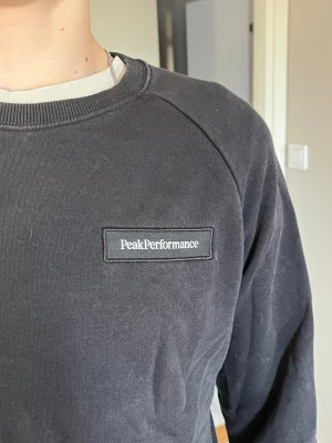 Svart Peak Performance sweatshirt - Svart sweatshirt från Peak Performance med klassisk rund hals och ribbade muddar. Diskret logotyp på bröstet. Tröjan har lång ärm och är gjord i mjuk bomull, perfekt för chill dagar eller när du vill ha en clean look. Storlek 160