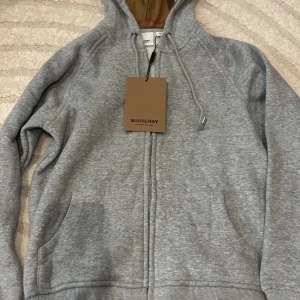 Grå hoodie från Burberry med huva - Snygg grå hoodie från Burberry i storlek M. Tröjan har dragkedja, snörning vid huvan och klassiskt Burberry-mönster på insidan av huvan. Tillverkad i mjuk bomull och har långa ärmar samt ribbade muddar. Perfekt för chill dagar och streetstyle inte äkta kommer med tagg osv.