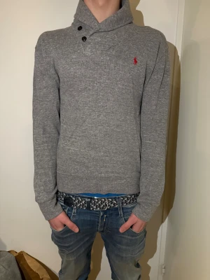 Grå tröja från Polo Ralph Lauren - Snygg grå tröja från Polo Ralph Lauren med klassisk röd logobrodyr på bröstet. Tröjan har lång ärm, ribbade muddar och en cool krage med två svarta knappar. Perfekt för dig som gillar stilrena och tidlösa plagg.