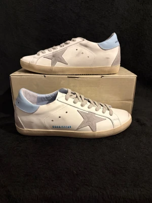 Golden Goose Superstar sneakers vit/blå - Golden Goose Superstar sneakers i vitt skinn med ljusblå häl och grå mockadetaljer. Klassisk stjärna på sidan, beige sula och snörning framtill. Snygg och stilren modell med coola kontraster och märkets logga på hälen. Perfekta för dig som gillar exklusiva och trendiga sneakers.