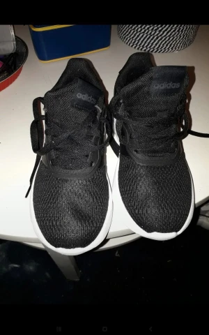 Svarta Adidas sneakers med guld och vit sula - Snygga svarta Adidas sneakers med vit sula och gulddetaljer på hälen. Ovandelen är i mesh-liknande syntetmaterial för bra andningsförmåga. Klassisk snörning och rund tå. Perfekta för dig som vill ha en sportig och stilren look.