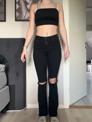 Svarta bootcut jeans med slitna knän - Säljer ett par svarta jeans med bootcut passform och snygga slitningar vid knäna. Hög midja och klassisk femficksdesign. Jeansen har en rak siluett upptill och lite utsvängda ben nertill, vilket ger en cool vibe. Perfekta för dig som gillar edgy stil.