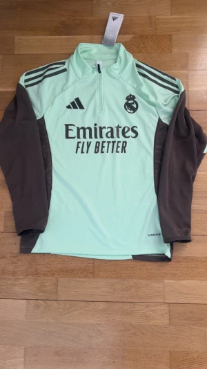 Real Madrid tracksuit  - Snygg Real Madrid träningshoodie från Adidas i mintgrönt och brunt med långärmade bruna ärmar och mintgrön kropp. Halv dragkedja framtill, klassiska Adidas-ränder på axlarna och Real Madrid-logga på bröstet. Perfekt för fotbollsträning och sport.