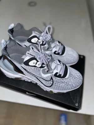 Nike React Element 55 D/MS/X sneakers - Nike React Element 55 D/MS/X sneakers i vit, grå och svart med mesh-överdel och coola detaljer. Sulan har ett vågigt mönster i vitt och olivgrönt, och skorna har snörning samt en tydlig Swoosh-logga på sidan. Perfekt för dig som gillar tech-inspirerad stil och komfort.