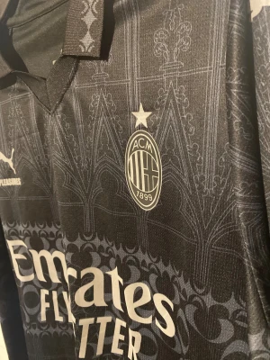 AC Milan x PUMA svart fotbollströja - AC Milan x PUMA matchtröja i svart med subtila grafiska detaljer inspirerade av gotisk arkitektur. Tröjan har klubbmärke, sponsorer och klassisk krage. Tillverkad i lätt och ventilerande funktionsmaterial, perfekt för fotboll eller streetwear. Nu under black friday veckan för endast 349 kr🔥🔥