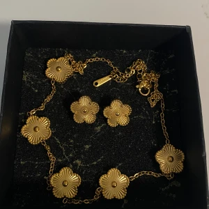 set i rostfritt stål halsband & örhängen - Snyggt set med halsband och örhängen i guldfärg, dekorerade med detaljerade blommor. Kedjan är tunn och smidig, och blommorna har en cool, räfflad yta som ger extra glans. Perfekt för dig som gillar unika accessoarer med blommig touch. Ask ingår ej