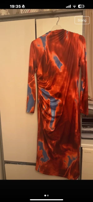 Röd och blå långärmad klänning - Säljer en långklänning med coolt abstrakt mönster i rött och blått. Klänningen har lång ärm och är figurnära med snygga rynkade detaljer på sidan. Materialet är glansigt och känns mjukt mot huden, perfekt för dig som gillar att sticka ut.