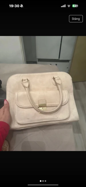 Beige handväska med gulddetalj - Stilren beige handväska i skinnimitation med två handtag och praktisk framficka med guldfärgat lås. Väskan har flera fack och dragkedja upptill, perfekt för att organisera dina grejer. Enkel och klassisk design som funkar till många outfits.