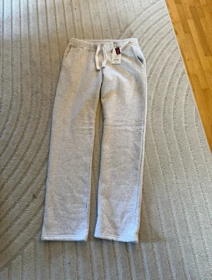 Ljusgrå mjukisbyxor från Russemerch - Sprillans nya jusgrå mjukisbyxor från Russemerch storlek M, med normal passform och snörning i midjan. Byxorna har raka ben och är tillverkade i mjukt bomullsmaterial, perfekta för chill dagar. Klassisk design med fickor på sidorna och elastisk midja.