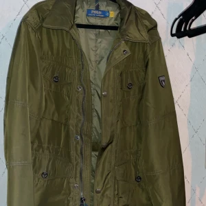 Olivgrön jacka från Polo Ralph Lauren - Säljer en snygg olivgrön jacka från Polo Ralph Lauren. Jackan har en dragkedja och knappar framtill samt ett stilrent märke på ärmen. Jackan håller bra värme i upp till 0° 