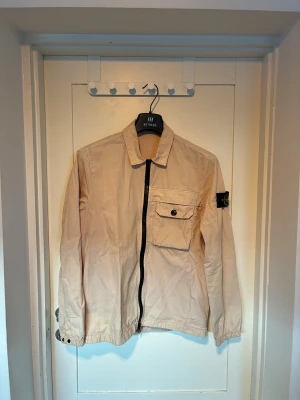 Beige overshirt från Stone Island - Snygg beige overshirt från Stone Island med klassisk krage, svart dragkedja och bröstficka med knapp. Ikonisk patch på ärmen. Perfekt för lager-på-lager och streetwear-vibe.