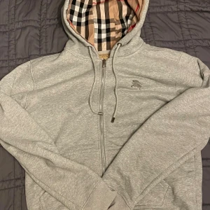 Grå Burberry hoodie med rutigt foder - Säljer en grå hoodie från Burberry med klassiskt rutigt foder i huvan och dragkedja framtill. Tröjan har broderad logga på bröstet, snörning i huvan och ribbade muddar. Perfekt för dig som gillar stilrena detaljer och ikoniskt mönster.