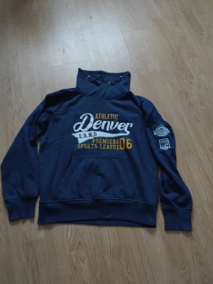 Mörkblå hoodie Denver Red & Blue - Snygg mörkblå hoodie från Red & Blue med tryck 'Denver Athletic' i vitt och gult på bröstet. Tröjan har snörning i huvan, känguruficka och coola patchar på ena ärmen. Perfekt för dig som gillar sportig streetstyle.