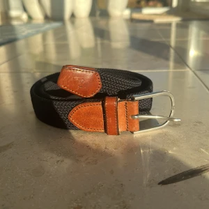 Marinblått flätat bälte  - Snyggt marinblått flätat bälte med bruna skinn-detaljer vid spännet och ändarna. Bältet har ett klassiskt silverfärgat metallspänne och är perfekt för att ge din outfit en stilren touch. Flätad design gör det flexibelt och bekvämt att bära. Priset är  diskuterbart!