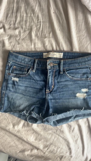 2000s talet blå jeansshorts Abercrombie & Fitch - Skit snygga vintage blå jeansshorts från Abercrombie & Fitch med slitna detaljer och fransig kant. Klassisk femficksmodell med knapp och dragkedja framtill. Söker då de inte passar längre men sitter verkligen så fint annars! 26W och 33L.