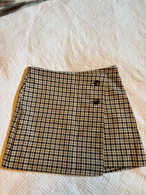 Rutig kortkjol från H&M med knappar - Superfin rutig kortkjol från H&M i svart, beige och brunt. Kjolen har två dekorativa knappar framtill och klassiskt rutmönster. 