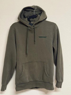 Olivgrön hoodie Peak Performance S - Olivgrön hoodie från Peak Performance i storlek S. Tröjan har huva med snörning, magficka och broderad logga på bröstet. Mjuk bomullskvalitet och ribbade muddar vid ärmslut och nederkant. Perfekt för chill eller träning.