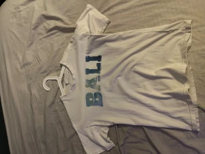 Vit t-shirt med Bali och Surfer-tryck - Vit t-shirt från Lindex med coolt tryck på framsidan. Ena sidan har texten 'BALI' i blå toner, andra sidan har ett surfmotiv med texten 'SURFER' i orange. Klassisk rund hals och korta ärmar. Perfekt för dig som gillar strandvibbar och avslappnad stil.