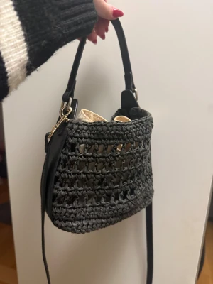 Svart virkad axelväska med skinnrem - Snygg svart axelväska med virkad design och detaljer i skinn. Väskan har en rundad form, guldfärgade metalldetaljer och en beige innerpåse. Perfekt storlek för mobilen och småsaker, med både handtag och axelrem för olika stilar.