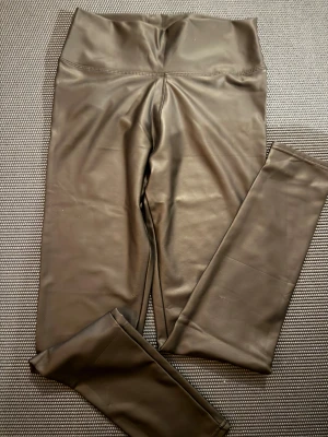 Svarta höga leggings i skinnimitation - Snygga svarta leggings med hög midja i skinnimitation. De har en tight passform och slät yta som ger en cool look. Perfekta att styla med oversized hoodie eller crop top. Materialet är syntet och ger en glansig finish.