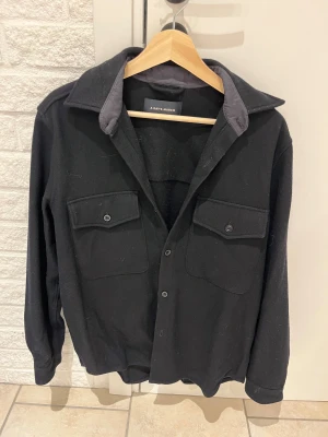 Mörkblå overshirt från A Day's March - Stilren svart overshirt från A Day's March med klassisk krage, två bröstfickor och knappar varav den knappen längst ner har tyvvär trillat av. Bortsätt från den är jackan i mycket bra skick. Tillverkad i ett tjockare ullmaterial som ger en cool och avslappnad look. Perfekt att bära lager på lager under kyligare dagar.