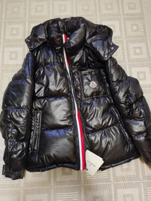 Svart Moncler pufferjacka med huva - Svart pufferjacka från Moncler med glansig finish, huva och ikonisk logga på bröstfickan. Jackan har en röd, vit och blå rand längs dragkedjan, quiltad design och innerfoder med serietidningsmönster. Perfekt för dig som vill ha en snygg och varm vinterjacka.
