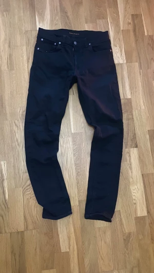 Svarta slim jeans från Nudie Jeans  - Snygga svarta jeans från Nudie Jeans Co med klassisk femficksdesign och dragkedja. Jeansen har en rak passform och är tillverkade i slitstark bomull. Perfekta för dig som gillar stilrena och tidlösa jeans med cool vibe. Har ett hål i vänster ficka, har även en fläck pp en av bakfickorna, bild kan fås om man frågar! Tveka inte på att skriva! 