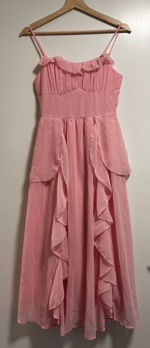 Lång rosa volangklänning med smala axelband - Supersöt långklänning i ljusrosa med smala axelband och volangdetaljer framtill. Klänningen har rynkade partier upptill och ett flowigt fall i kjolen. Perfekt för dig som vill ha en romantisk och feminin look.