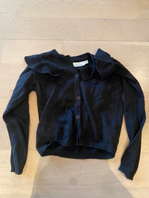 Svart kofta med volang från KappAhl - Svart kofta från KappAhl med glittriga inslag och snygga volanger på axlarna. Koftan har lång ärm, knappar framtill och ribbade muddar vid ärmslut och nederkant. Perfekt för dig som gillar lite extra detaljer på plaggen.