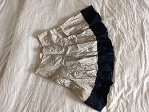 Beige och marinblå kjol från STOCKH LM - Snygg beige kjol med bred marinblå volangkant från STOCKH LM. Kjolen är medellång och har veckad design.. Tillverkad i bomull och acetat för en lätt och luftig känsla. Perfekt för dig som gillar en stilren men ändå lekfull look.