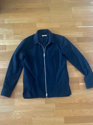 Svart overshirt med dragkedja J.Lindeberg - Stilren svart overshirt med klassisk krage och hel dragkedja framtill. Jackan har lång ärm och en diskret bröstficka. Perfekt för lager på lager och enkel att matcha med olika outfits.