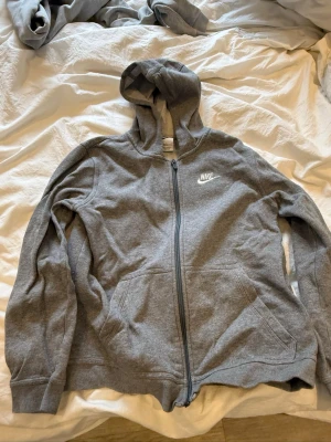 Grå zip-hoodie från Nike - Klassisk grå hoodie från Nike med dragkedja framtill och vit logga på bröstet. Tröjan har huva med dragsko och två stora fickor framtill. Tillverkad i mjukt sweatshirtmaterial som är skönt att ha på sig. 