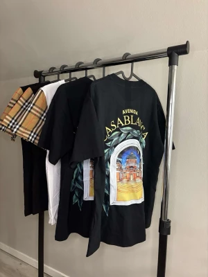 Svart Casablanca Avenida t-shirt - Säljer en svart t-shirt från Casablanca med stor färgglad grafisk print på ryggen där det står 'Avenida Casablanca' och ett fönster med arkitekturmotiv. T-shirten har normal passform och korta ärmar. Perfekt för dig som gillar streetwear och unika prints.
