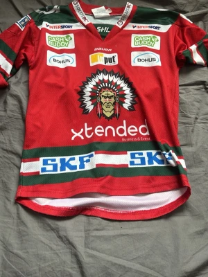 Frölunda Indians matchtröja röd - Röd Frölunda Indians hockeytröja från Bauer med gröna och vita detaljer. Stora tryckta loggor och sponsormärken på både fram- och baksida. Klassisk v-ringning och klubbens ikoniska indianmotiv på bröstet. Perfekt för hockeyfans!