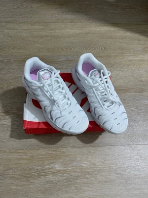 Nike Air Max Plus vita och rosa sneakers - Nike Air Max Plus sneakers i vitt meshmaterial med rosa och grå detaljer. Skorna har synlig Air Max-dämpning i sulan, vågiga linjer på sidan och klassisk Nike Swoosh i rosa. Låg modell med vita skosnören och rosa insida. Perfekta för dig som gillar sportig och trendig stil.