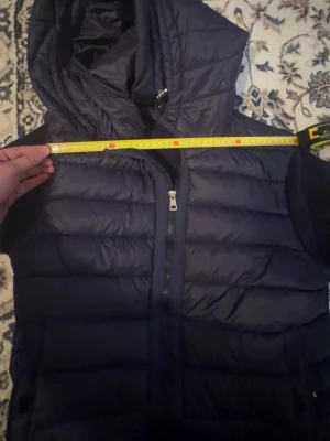 Mörkblå dunväst från Moncler - Snygg mörkblå dunväst från Moncler med huva och dragkedja framtill. Västen har quiltad design och Moncler-logga på ärmen. Perfekt för lager-på-lager och håller dig varm under kyliga dagar. Klassisk look med sportig touch.