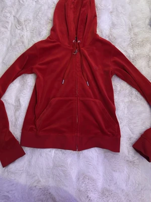 Röd velourhoodie från Juicy Couture - Juicy Couture hoodie i röd velour med dragkedja framtill, stora fickor och justerbar huva med snören. Klassisk logga på dragkedjan och mjukt material som ger en lyxig känsla. Perfekt för en avslappnad och trendig look.