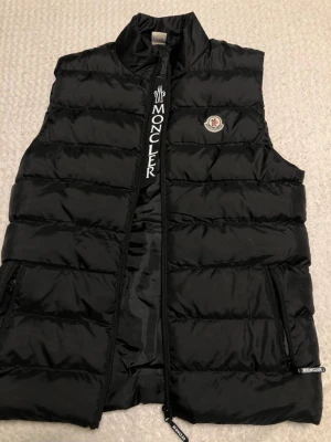 Svart Moncler pufferväst strl L - Säljer en svart pufferväst från Moncler i storlek L. Västen har hög krage, broderad Moncler-logga på bröstet och dragkedja framtill med tydlig Moncler-text. Tillverkad i blank polyester och perfekt för lager-på-lager-stil.