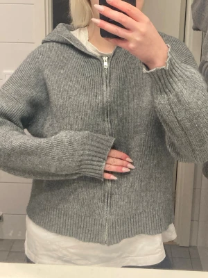 Grå stickad hoodie med dragkedja - Mysig grå stickad hoodie med dragkedja framtill och luva. Tröjan har ribbstickade muddar och en avslappnad passform. Perfekt för lager på lager och chill dagar.