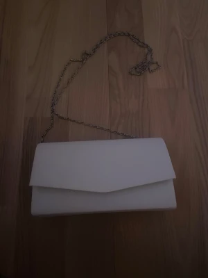 Vit kuvertväska med kedjerem - Stilren vit kuvertväska med silverfärgad kedjerem och magnetlås. Väskan har en klassisk rektangulär form och är perfekt för att bära det viktigaste. Materialet ser ut att vara slätt konstläder. Enkel och snygg design som passar till många outfits. Kedjan går att ta loss.